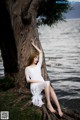 A woman in a white dress sitting on a tree by the water.